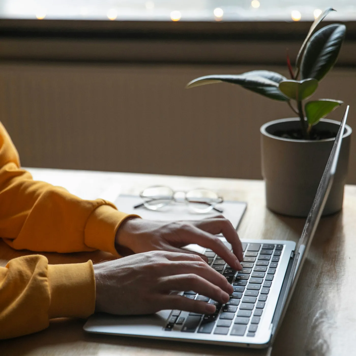 Mãos digitando num notebook sobre a mesa, com um par de óculos e um vaso de planta ao lado